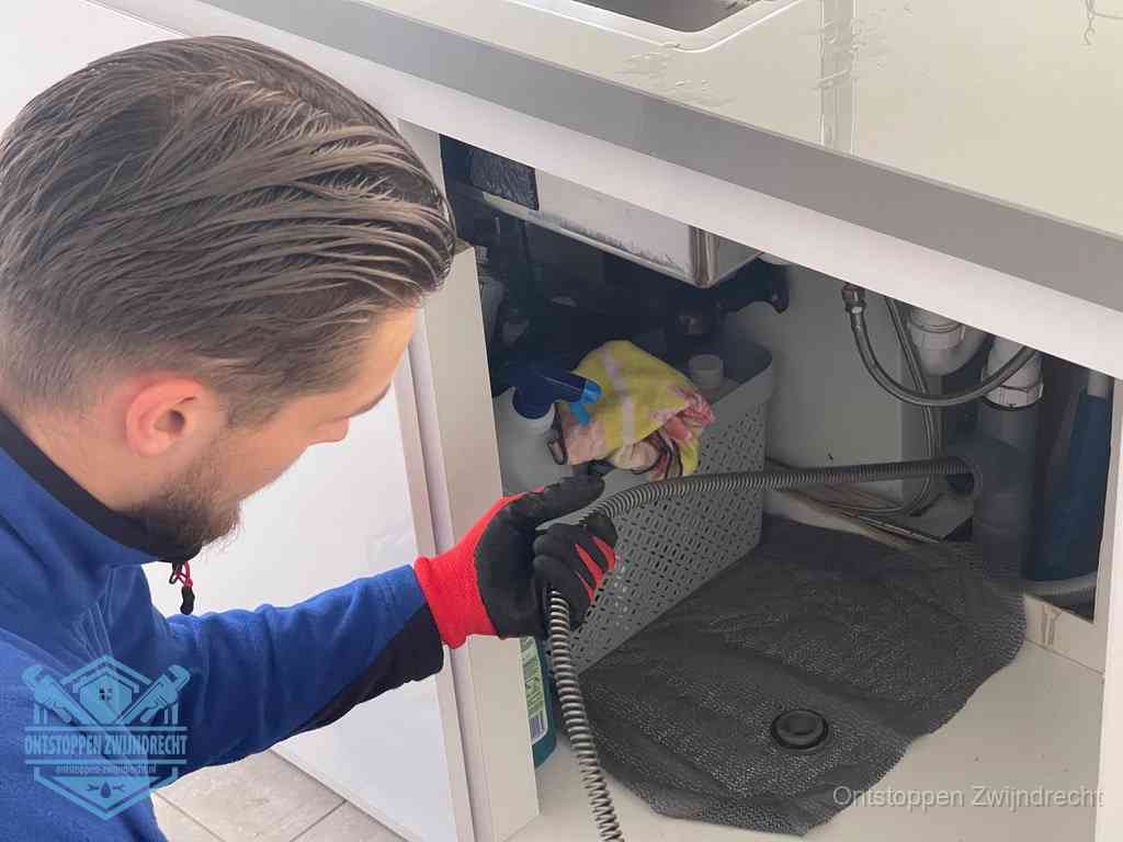 Een monteur voert een flexibele ontstoppingsveer in onder het keukenkastje, langs de sifon, om de gootsteen te ontstoppen in Zwijndrecht