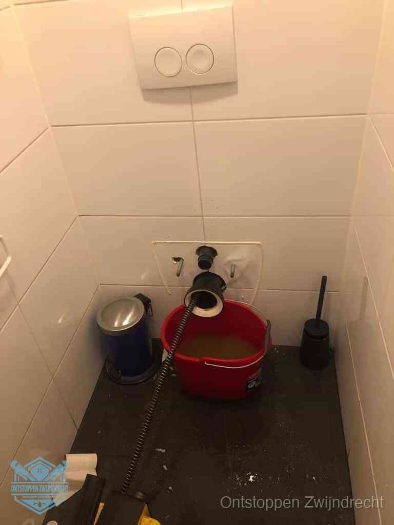 Een ontstopte wand-wc met verwijderde toiletpot waar een ontstoppingsveer in de wanduitgang verdwijnt, het achterhalen waar de verstopping zit in Zwijndrecht