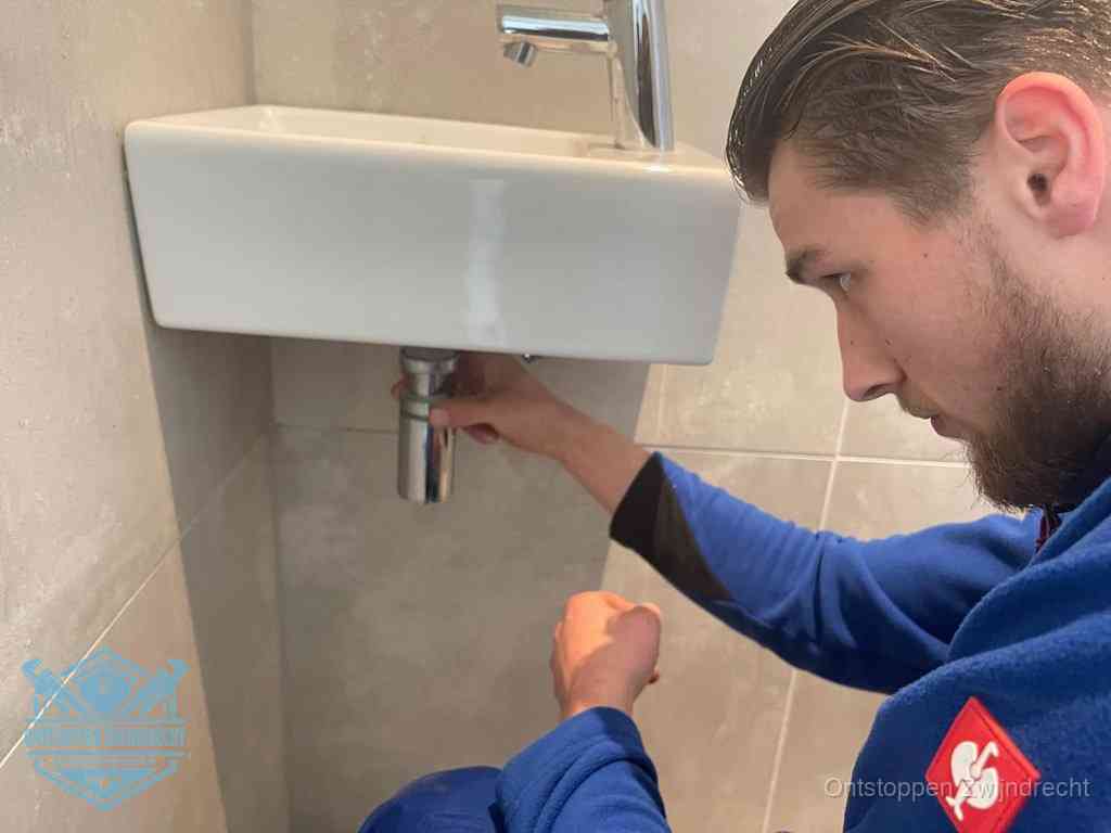 Een monteur draait met de hand de zwanenhalssifon onder een fonteinwasbak in de toiletruimte los om de bron van rioollucht in Zwijndrecht te vinden