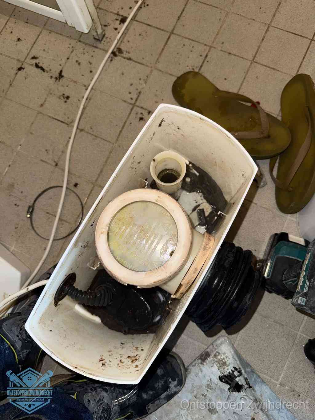 Een gedemonteerde toiletpot op de vloer met ontstoppingsveer en gereedschap ernaast, het verwijderen van een vastzittend voorwerp uit de wc in Zwijndrecht
