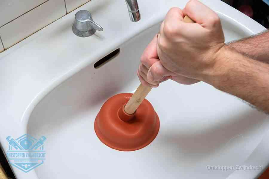 Twee handen gebruiken een rode rubberen plopper op een sanitair-afvoer om het toilet te ontstoppen in Zwijndrecht