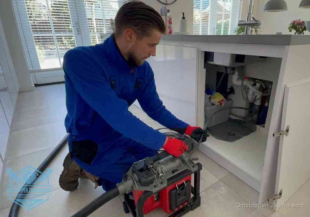 Een monteur voert een professionele elektrische ontstoppingsmachine in de afvoer onder de spoelbak tijdens het gootsteen ontstoppen in Zwijndrecht